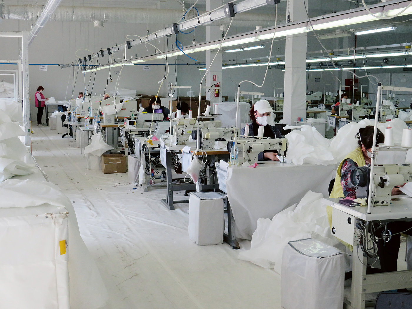 Foto: In einer großen Halle sitzen Personen hinter Industrienähmaschinen. Sie nähen mit weißem Garn ein weißes Gewebe.
