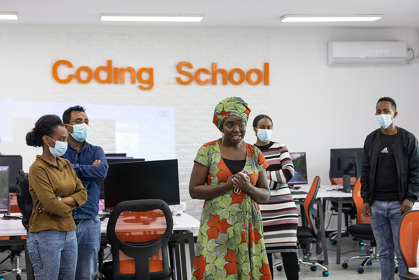 Foto: Eine Frau steht in der Mitte eines Raumes und spricht. Kreisförmig stehen weitere Personen um sie herum; alle tragen eine OP-Maske. Im Hintergrund der Schriftzug „Coding School“ an der Wand.