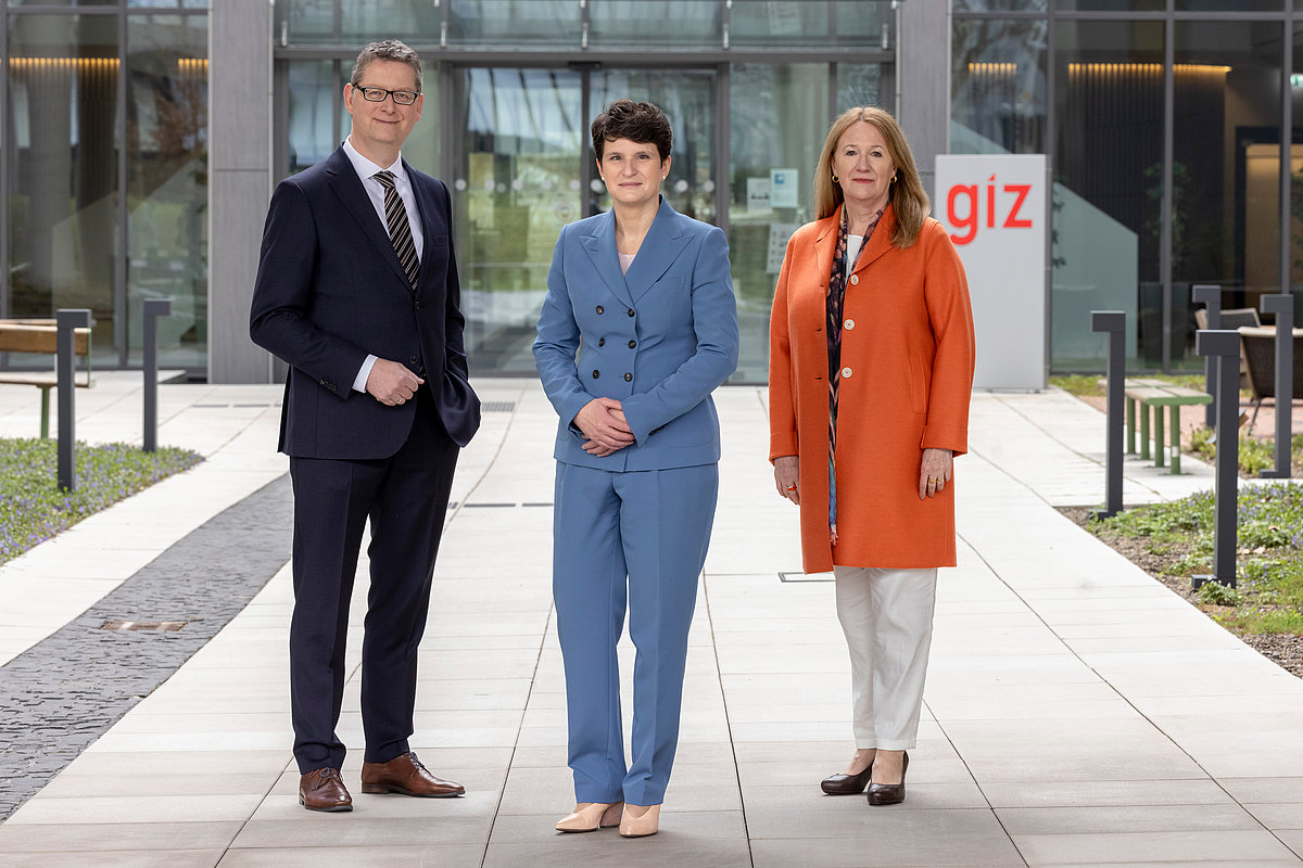 Foto: Drei Personen stehen, leicht zueinander versetzt, nebeneinander auf einem Weg, der zu einem Gebäude der GIZ führt.