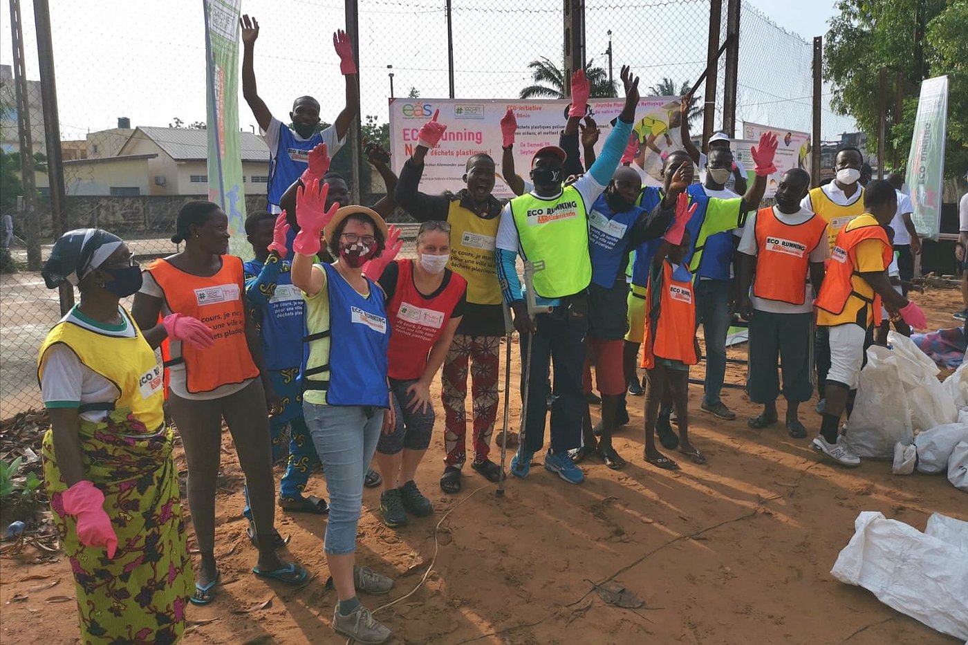 Foto: Personen mit Mund-Nase-Bedeckung posieren vor einem Schild mit der Beschriftung „ECO-running – Non aux déchets plastiques!“. Sie tragen Westen mit unterschiedlichen Farben.