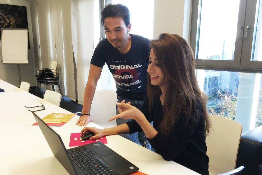 Foto: Zwei Personen vor einem Tisch mit einem geöffneten Laptop. Eine Person sitzt davor und bedient das Gerät, eine weitere Person steht daneben.