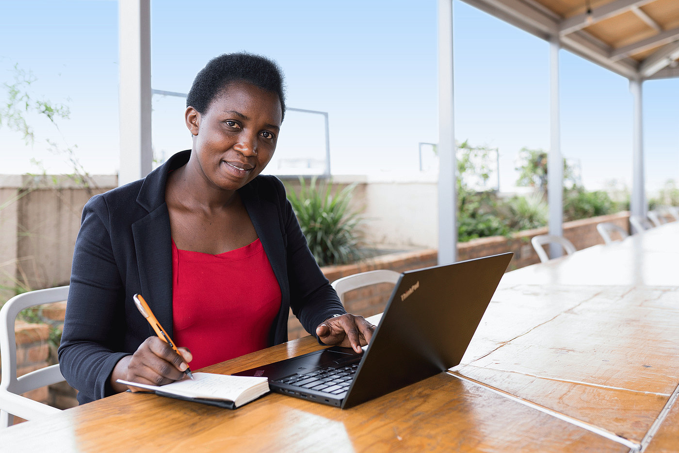 Foto: Eine Frau sitzt im Freien an einem Schreibtisch. Vor ihr steht ein geöffneter Laptop. Sie notiert etwas in ihrem Notizblock, welcher neben dem Laptop auf dem Tisch liegt.