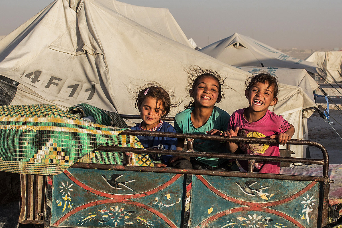 Foto: Drei Kinder sitzen erhöht auf einer Ladefläche mit Metallgitter. Sie lachen. Im Hintergrund große Zelte.