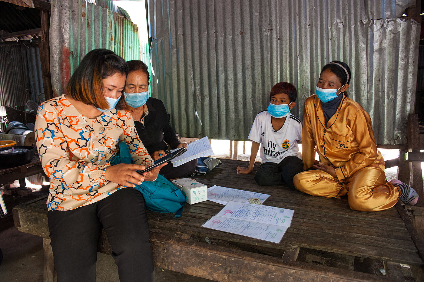 Foto: Mehrere Personen auf einer Holzplattform. Alle tragen eine OP-Maske.  Auf dem Boden einige Schriftstücke. Eine Person hält ein Tablet in den Händen, eine weitere Person blickt auf den dargestellten Inhalt.