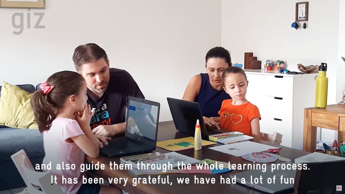 Foto: Zwei junge Mädchen sitzen an einem Tisch vor aufgeklappten Laptops. Ihre Eltern sitzen neben ihnen und schauen ihnen zu.