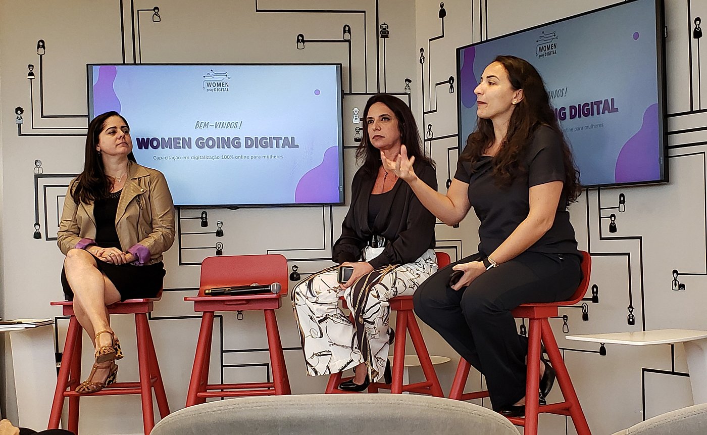 Foto: Drei Frauen sitzen auf erhöhten Stühlen. Im Hintergrund Monitore an den Wänden mit dem Titel „Women going digital“.