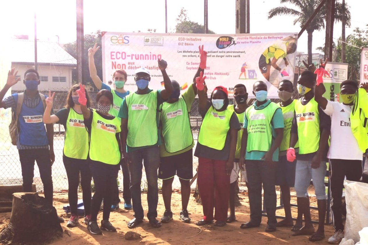 Foto: Personen mit Mund-Nase-Bedeckung posieren vor einem Schild mit der Beschriftung „ECO-running – Non aux déchets plastiques!“. Sie tragen vorwiegend Westen mit gelber/grüner Farbe.