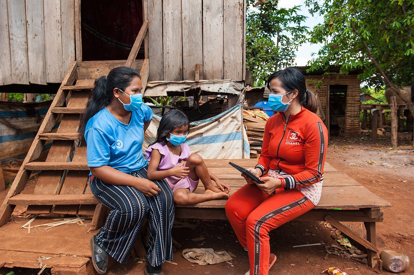 Foto: Zwei Frauen und ein Kind sitzen auf einer Holzplattform vor einem Holzhaus. Alle tragen eine OP-Maske. Eine Frau hält ein Tablet mit beiden Händen.