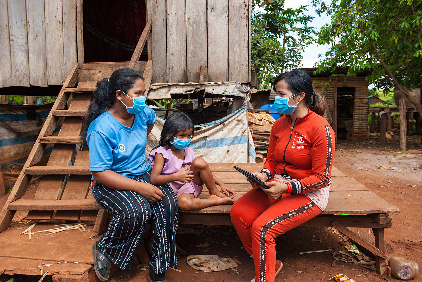 Foto: Zwei Frauen und ein Kind sitzen auf einer Holzplattform vor einem Holzhaus. Alle tragen eine OP-Maske. Eine Frau hält ein Tablet mit beiden Händen.