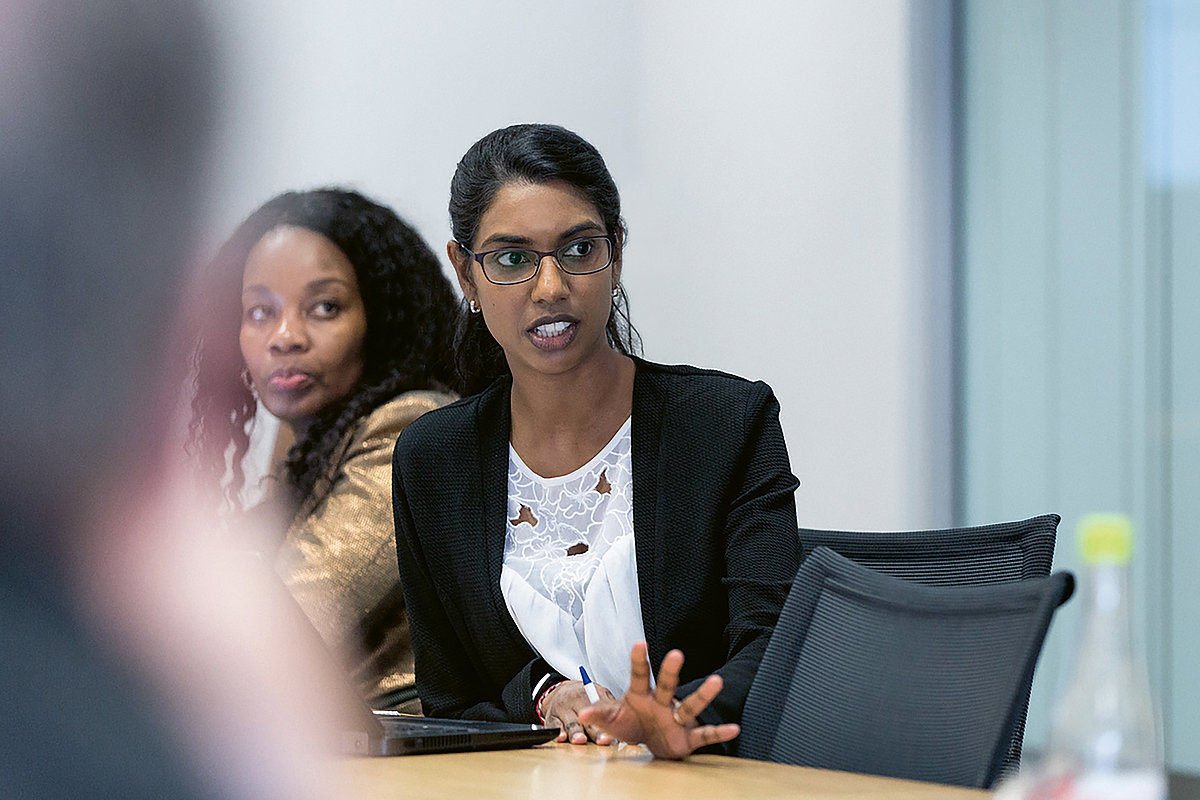 Foto: Zwei Frauen sitzen nebeneinander an einem Tisch. Eine Frau hat ein geöffnetes Notebook vor sich stehen während sie spricht.