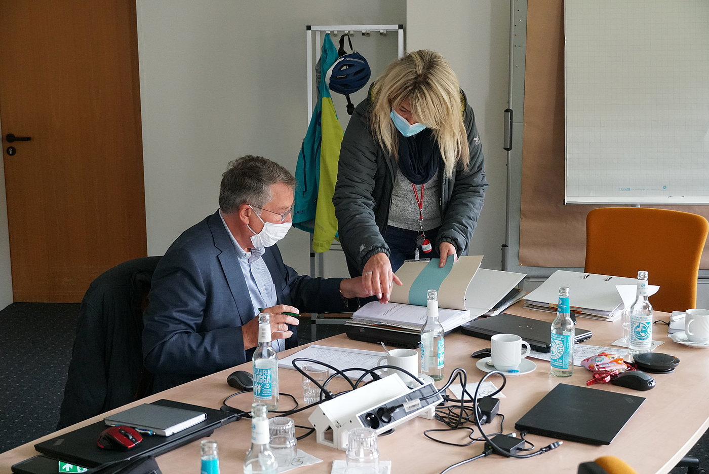 Foto: Ein Mann sitzt mit einer OP-Maske an einem Schreibtisch. Weitere Schreibtische stehen daneben; sie bilden zusammen eine größere Schreibtischfläche. Auf den Schreibtischen Anschlüsse und Geräte. Neben dem Mann steht eine Frau, welche ebenfalls eine OP-Maske trägt. Sie zeigt in einem Ordner auf eine Seite. Im Hintergrund ein Flipchart.