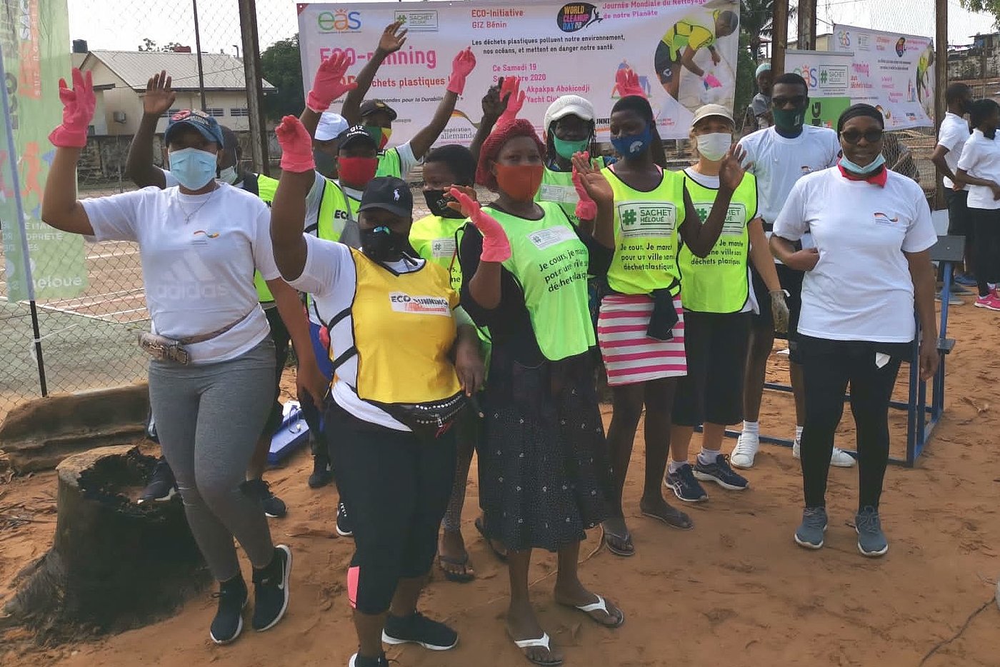 Foto: Personen mit Mund-Nase-Bedeckung posieren vor einem Schild mit der Beschriftung „ECO-running – Non aux déchets plastiques!“. Einige tragen Westen mit gelber/grüner Farbe.