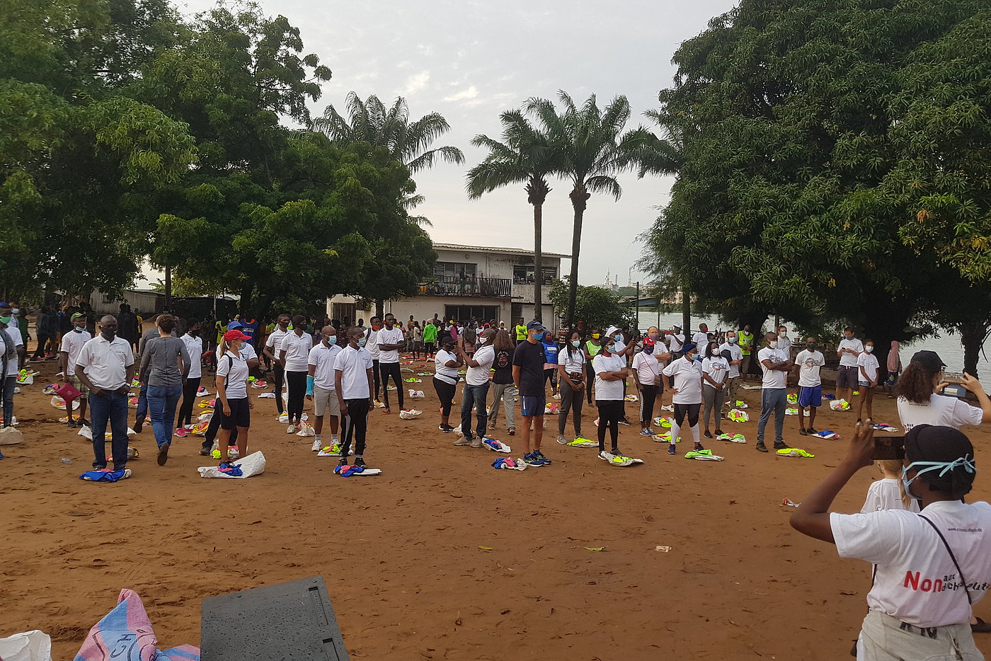 (2) Foto: Eine große Gruppe von Personen, die in Reihen neben- und hintereinander stehen. Alle tragen Mund-Nase-Bedeckungen. Auf dem Boden jeweils ein leerer Müllsack.