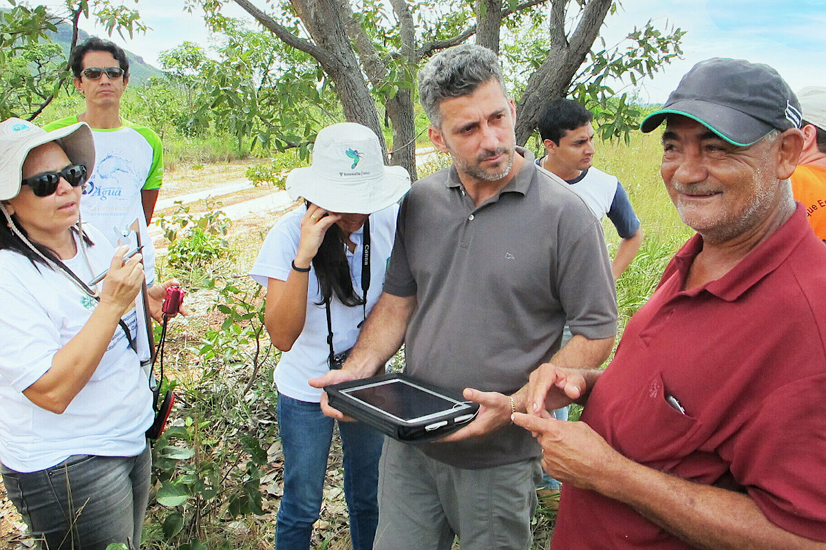 Foto: Mehrere Menschen stehen auf einem Feld vor einem Baum. Ein Mann hält ein Tablet in den Händen.
