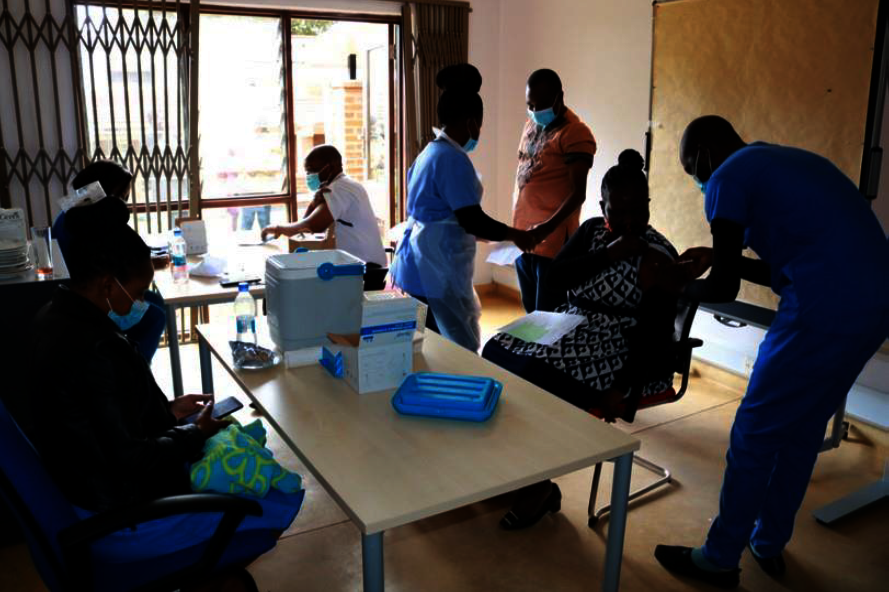 Foto: Verschiedene Menschen sitzen teilweise mit Mundschutz an Tischen. Eine Frau wird gerade geimpft.