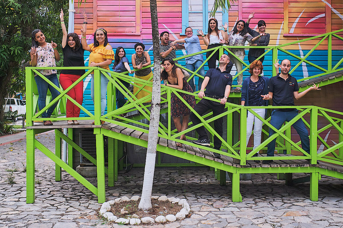 Foto: Mehrere Personen stehen auf einem Aufgang mit grünem Geländer vor einem bunten Gebäude.