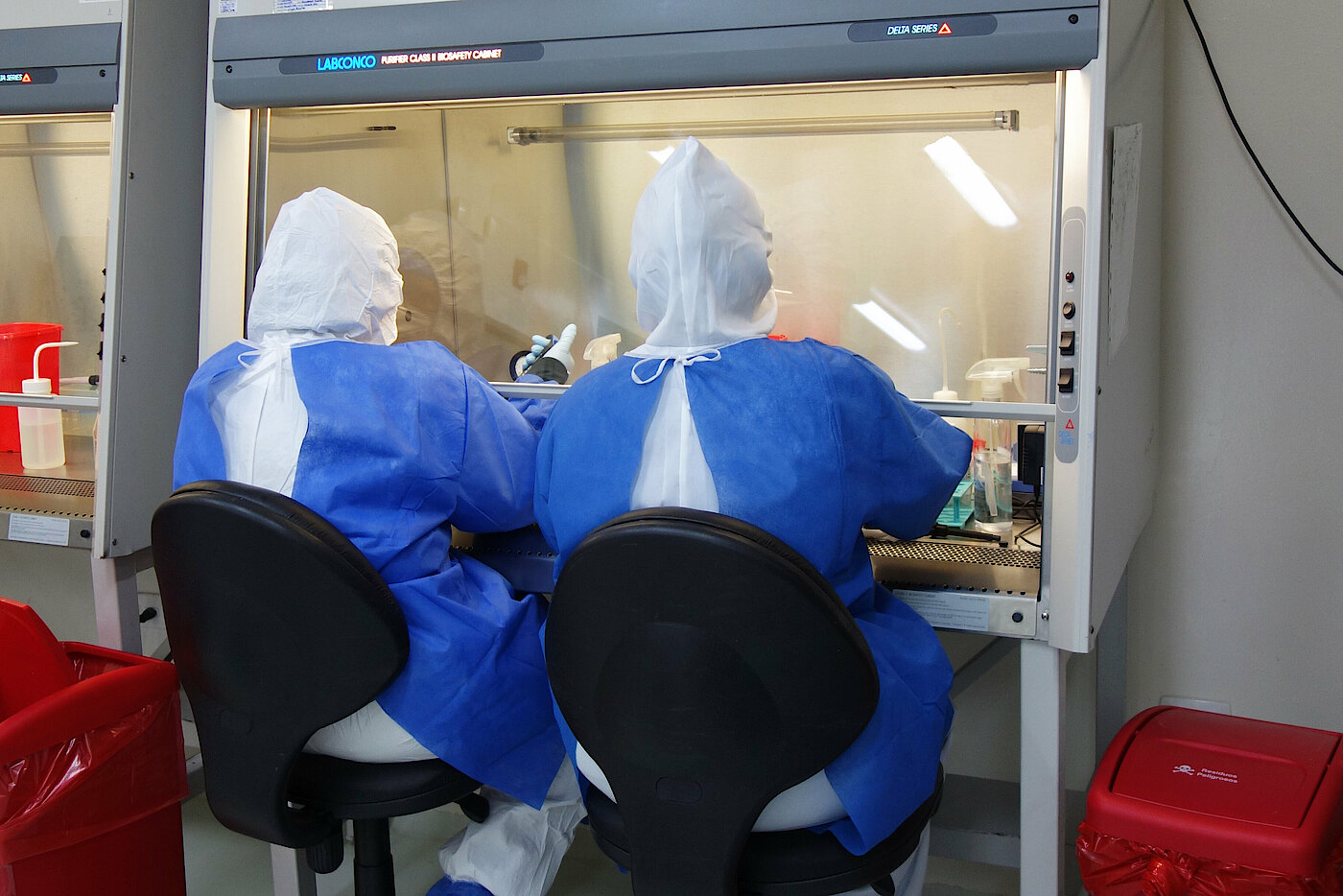 Foto: Zwei Personen in Schutzkleidung arbeiten mit ihren Händen in einem kleinen Labor mit Glasfront.