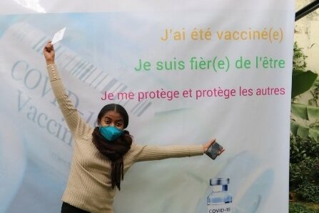 Foto: Eine junge Frau mit Mundschutz streckt die Arme hoch. Sie steht vor einem Plakat mit der Aufschrift: J’ai été vacciné(e). Je suis fièr(e) de l’être. Je me protège et protége les autres.