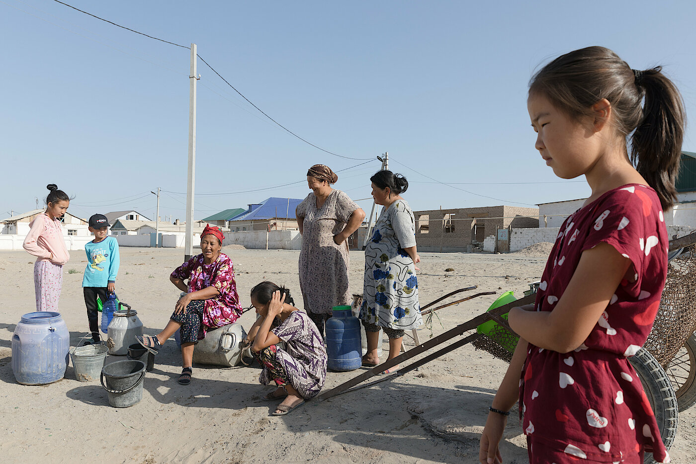 Frauen und Kinder stehen und sitzen neben leeren Eimern und Tonnen auf einem ausgetrockneten Boden.