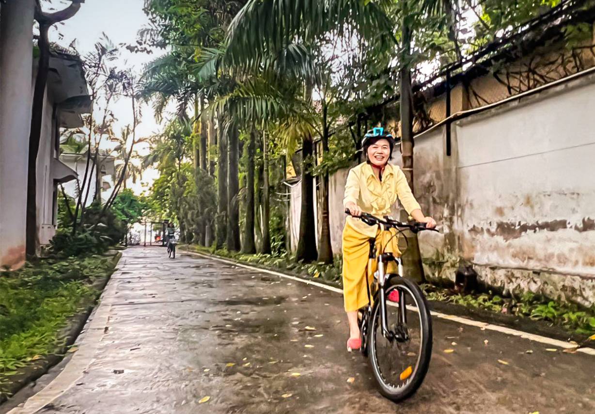 Foto: Eine Frau mit Helm sitzt auf einem Fahrrad, im Hintergrund sind am Wegesrand Palmen zu sehen.