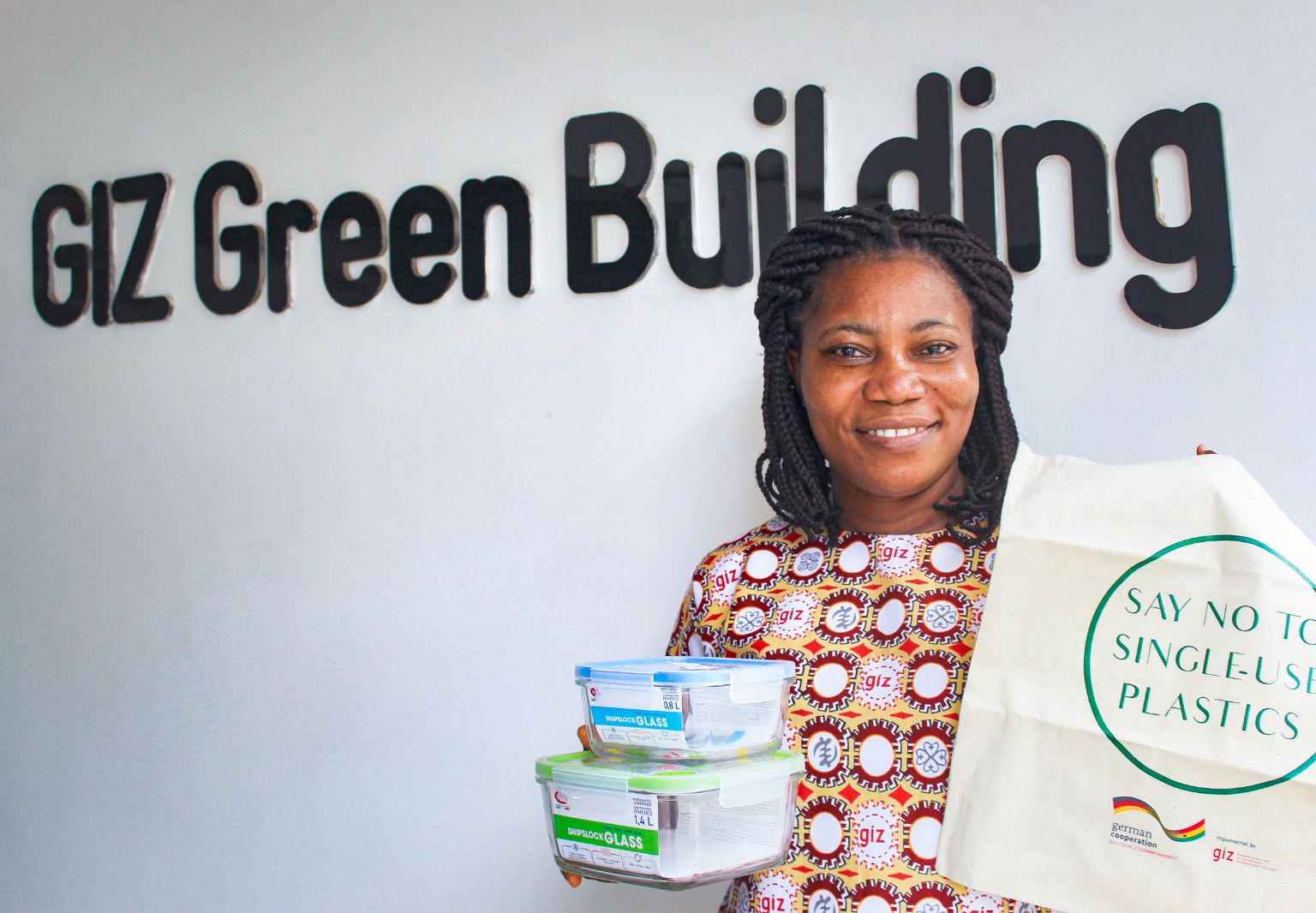 Foto: Eine Frau steht vor einer Wand mit der Aufschrift: „GIZ Green Building“. In den Händen hält sie zwei Glasboxen mit Deckel und eine Stofftasche mit der Aufschrift: „Say no to single-use plastics“.