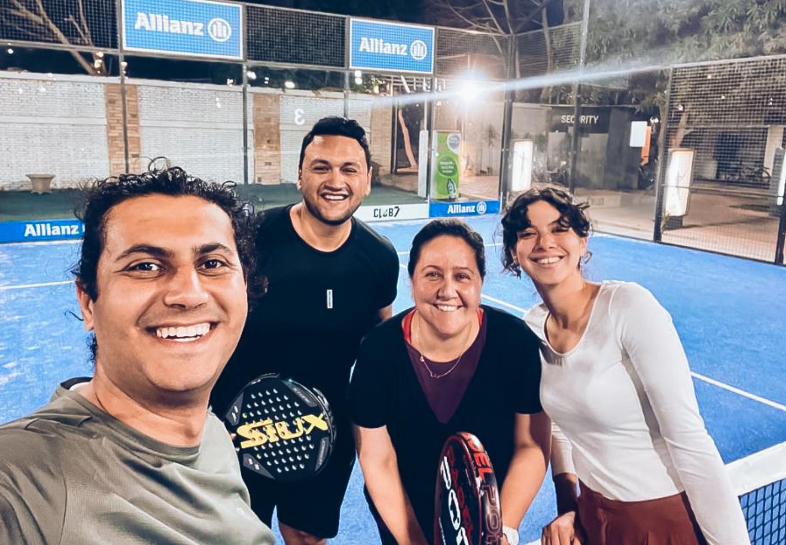 Foto: Vier Personen stehen teilweise mit Schlägern auf einem Tennisplatz und lächeln.