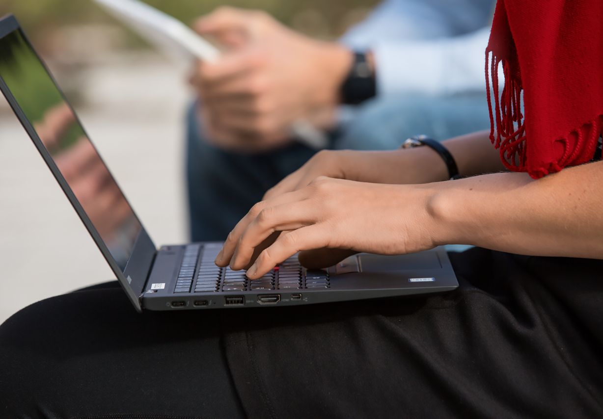 Foto: Eine Person tippt auf einem Laptop, das sie vor sich auf den Beinen abgestellt hat.