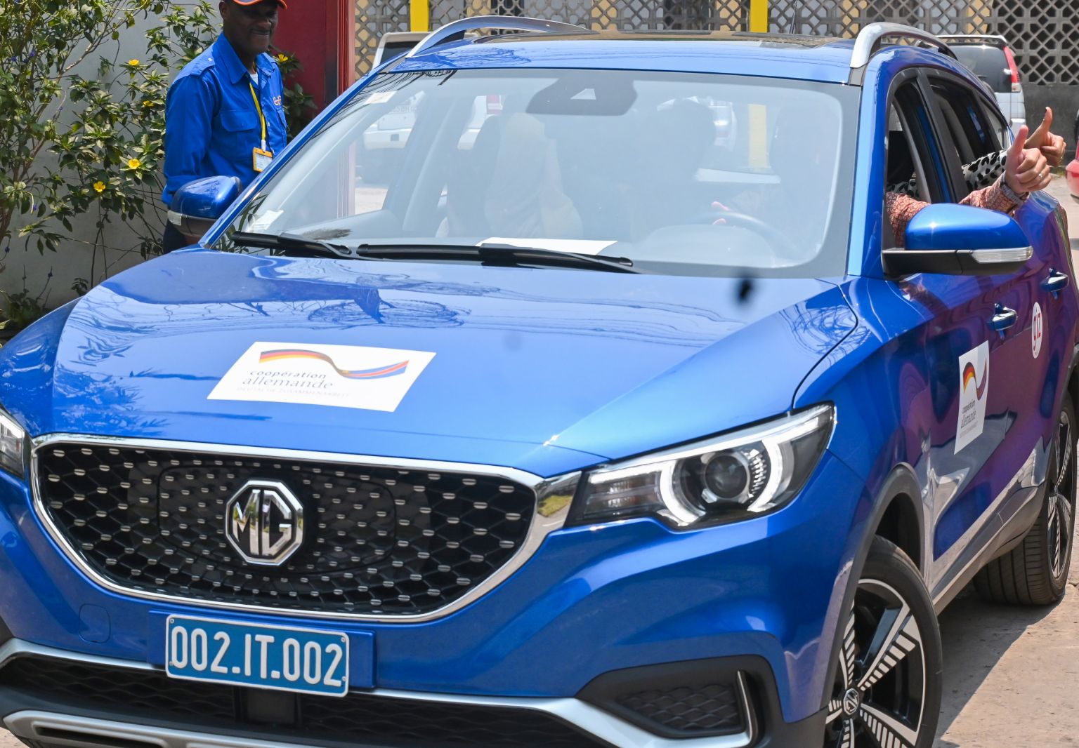 Foto: Ein blaues Auto, auf dessen Motorhaube und Türen Aufkleber mit dem deutsch-kongolesischen Kooperationslogo sowie dem GIZ-Logo zu sehen sind. Die Insassen des Autos strecken ihre gehobenen Daumen aus den geöffneten Autofenstern.