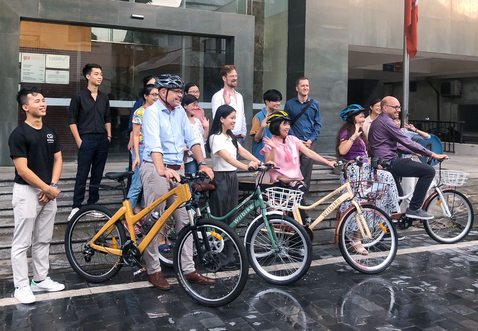 Foto: Eine Gruppe von Personen, einige von ihnen mit Fahrradhelmen, steht vor einem Gebäude mit GIZ-Logo. Alle lächeln, einige winken. Vier Personen halten ein Fahrrad in der Hand.