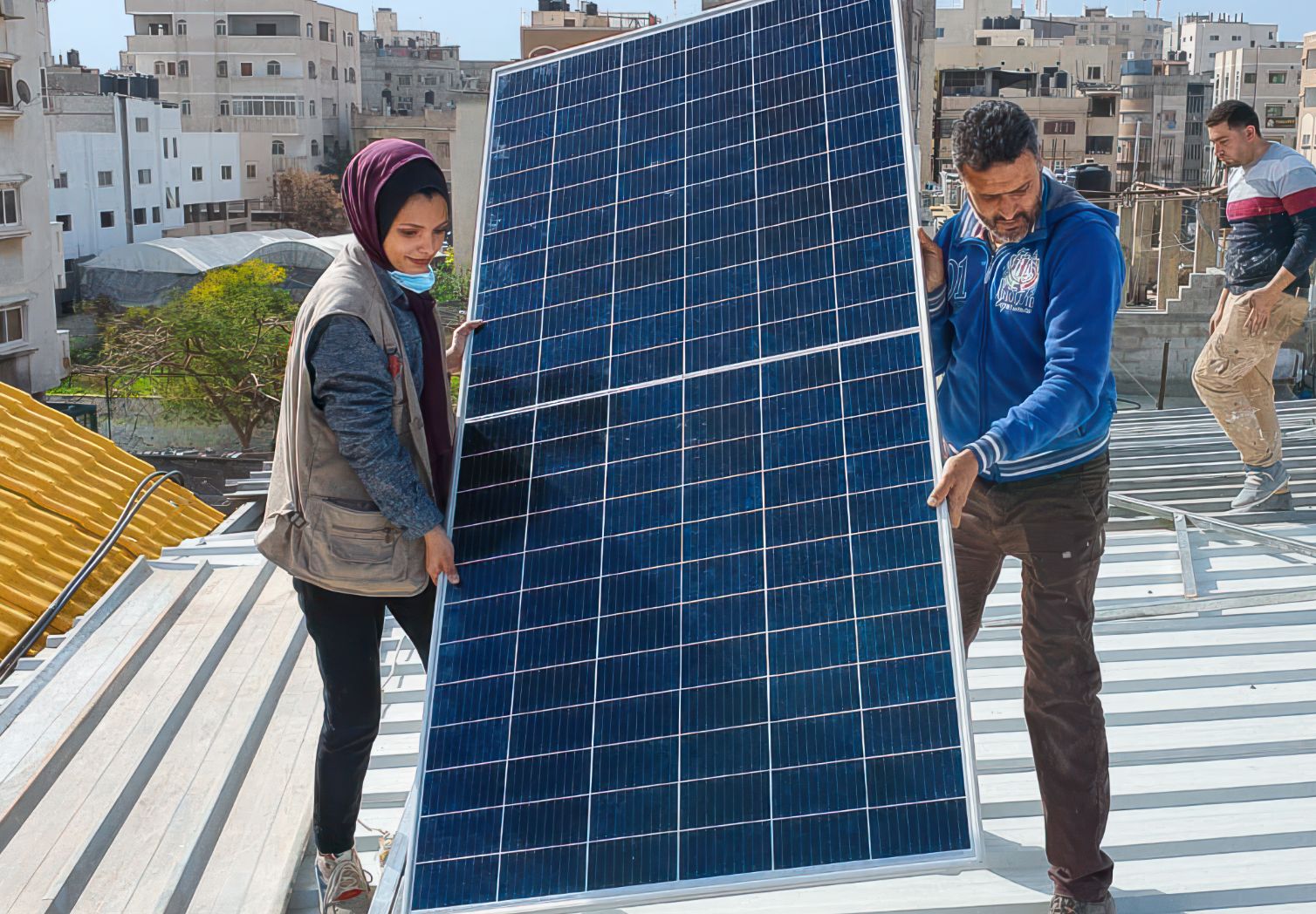 Foto: Eine Frau mit Kopftuch und ein Mann befinden sich auf dem Dach eines Hauses uns tragen gemeinsam ein großes Solarpaneel. 
