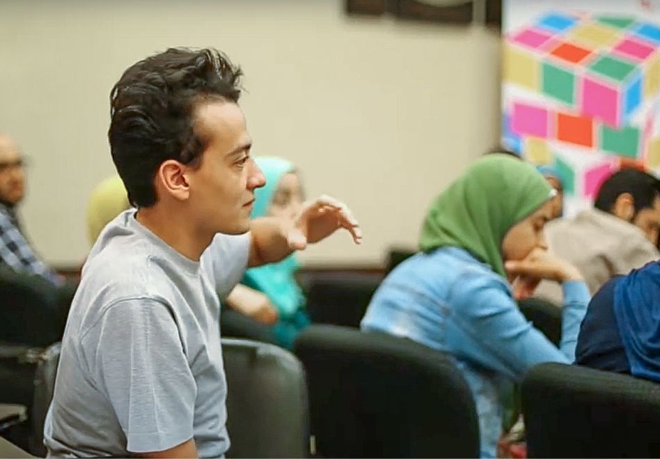 Foto: Ein junger Mann hebt in einem Klassenraum die Hand. Im Hintergrund sieht man weitere Lernende, die Frauen tragen Kopftücher.