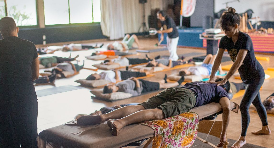 Foto: Ein Mann liegt auf dem Bauch auf einer Liege und bekommt eine Schultermassage. Im Hintergrund liegen Menschen auf Yogamatten.