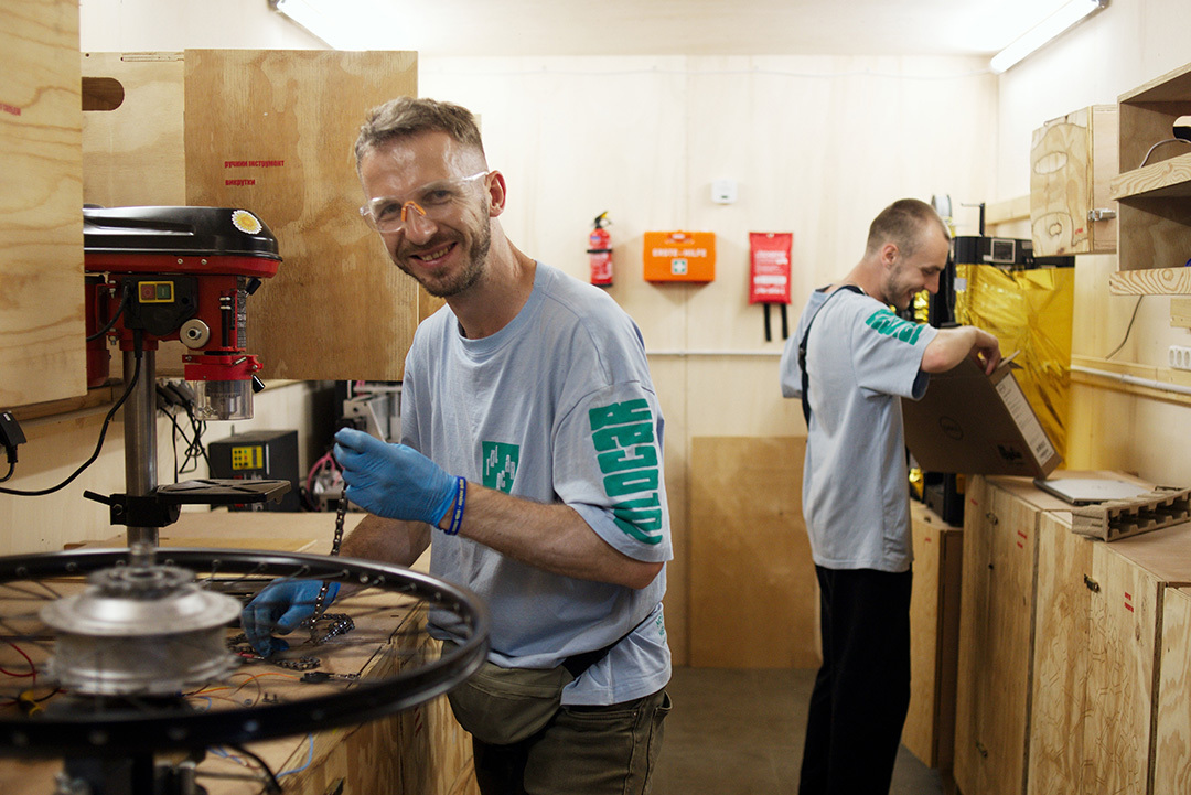 Ein Mann mit Schutzbrille repariert in einer Werkstatt eine Fahrradkette. Ein anderer Mann hat einen Aktenordner in der Hand. Beide lächeln. &copy; GIZ / Sergiy Bezborodko