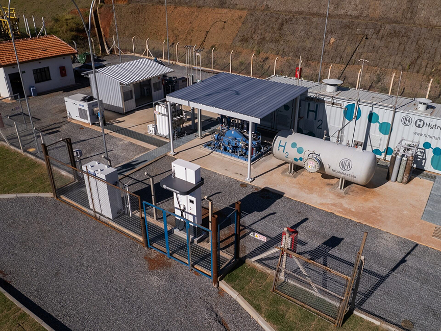 Eine eingezäunte Pilotanlage zur Produktion von grünem Wasserstoff steht am Rande eines Berghangs. Auf einem der verschiedenen, miteinander verbundenen Kessel ist die Aufschrift H2 zu lesen.