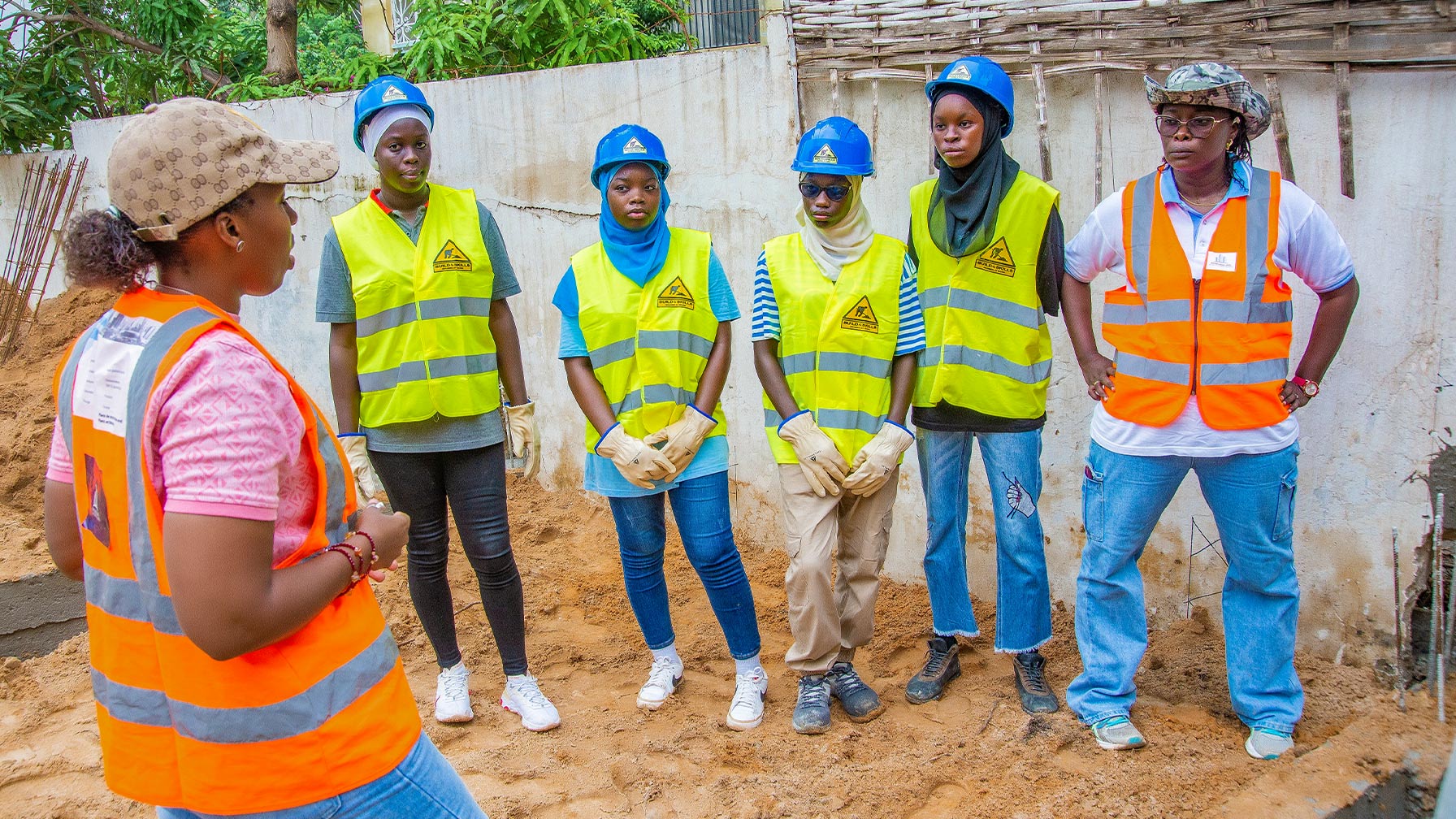 Gruppenfoto mit Djélia Diagne (links im Profil) im Gespräch mit fünf jungen Frauen in Schutzhelmen und Warnwesten auf einer Baustelle.