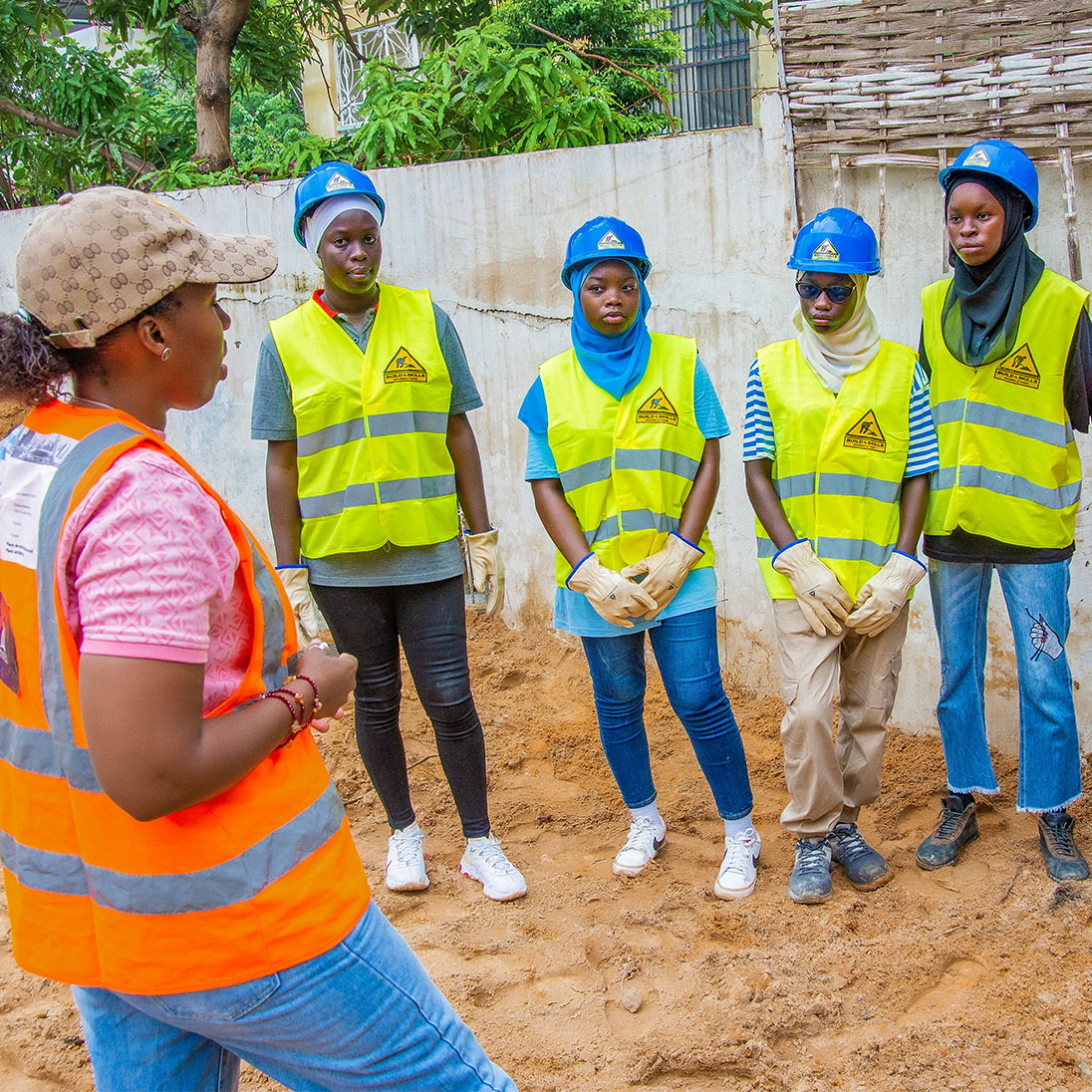 Gruppenfoto mit Djélia Diagne (links im Profil) im Gespräch mit fünf jungen Frauen in Schutzhelmen und Warnwesten auf einer Baustelle.