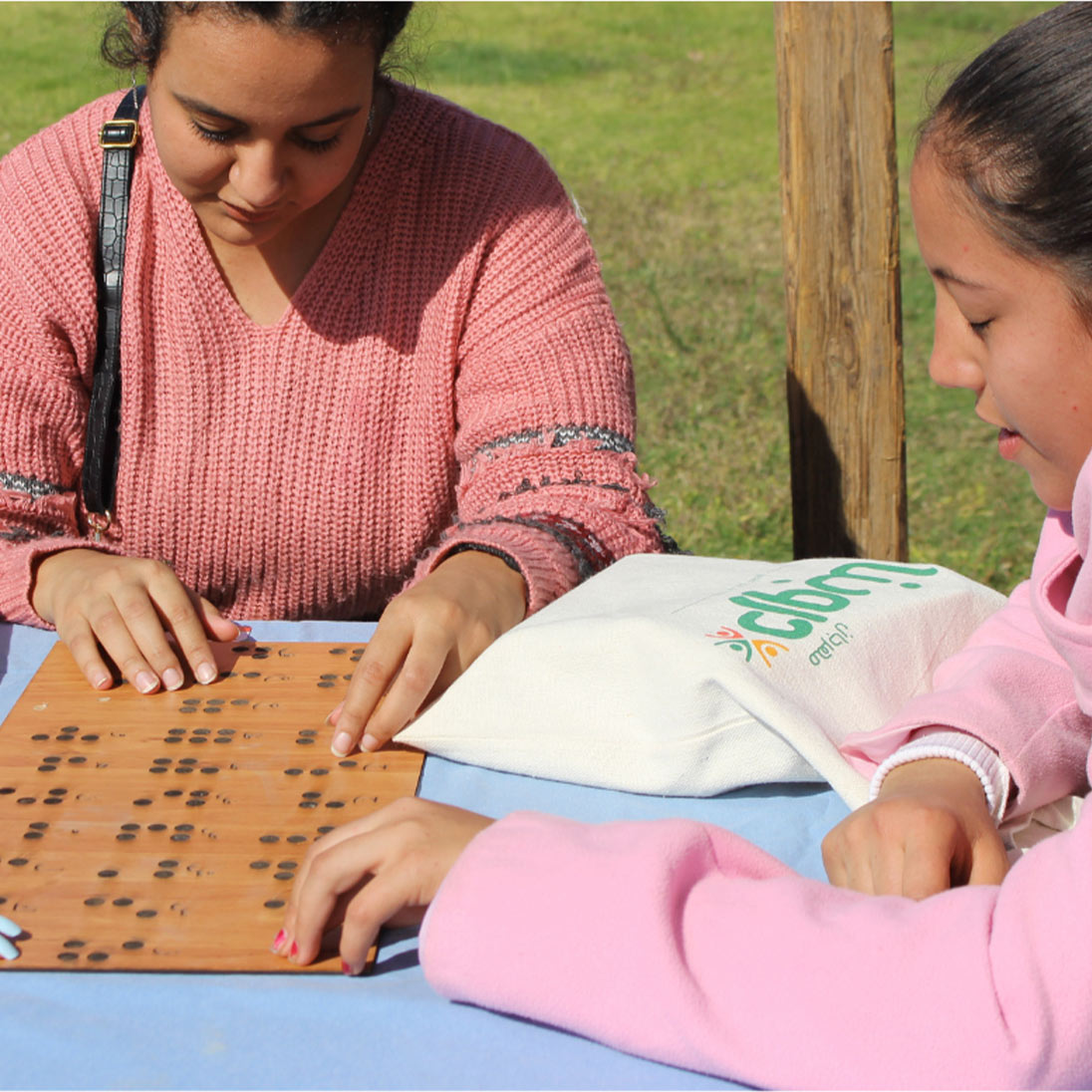 Drei junge Frauen sitzen an einem Tisch im Freien und ertasten mit den Fingern ein hölzernes Brett mit Brailleschrift.