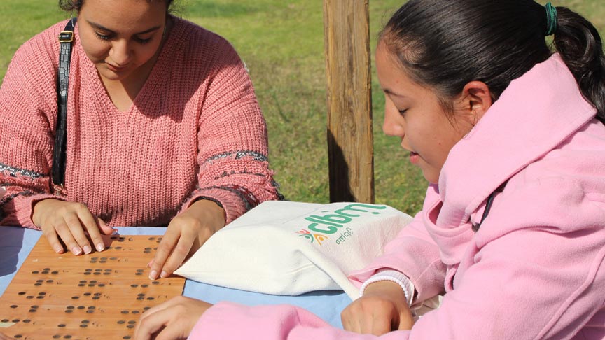 Drei junge Frauen sitzen an einem Tisch im Freien und ertasten mit den Fingern ein hölzernes Brett mit Brailleschrift.