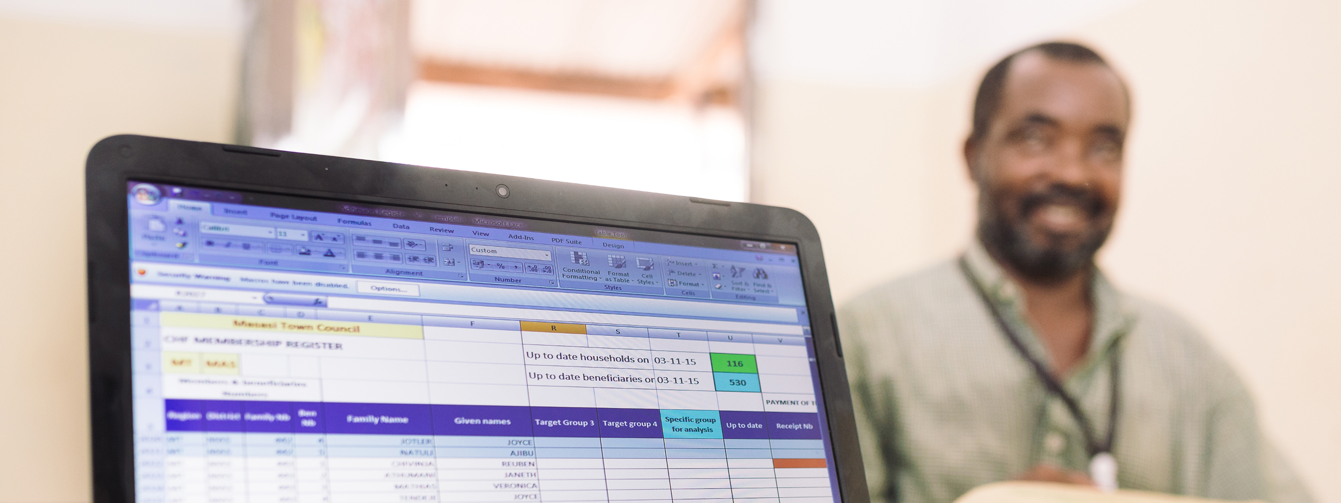 Ein Mensch tippt auf einem Laptop mit Excel-Tabelle, im Hintergrund sitzt ein lächelnder Mann an einem Schreibtisch.