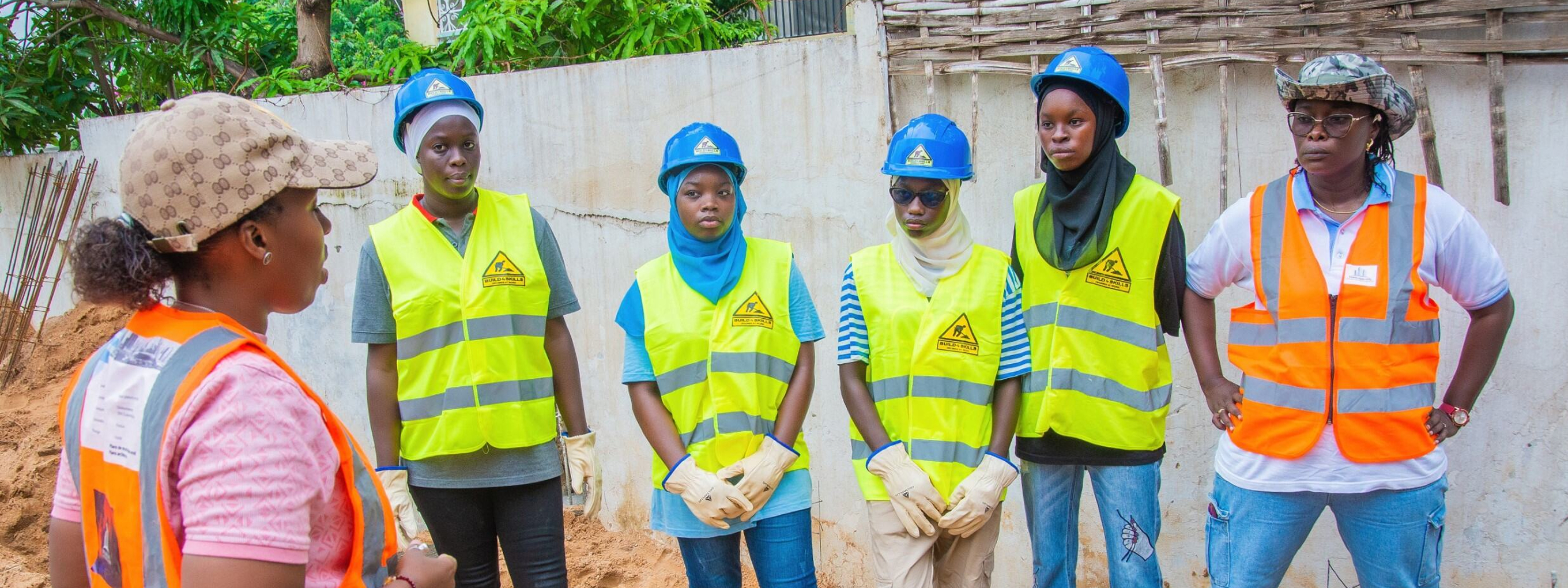 Gruppenfoto mit Djélia Diagne (links im Profil) im Gespräch mit fünf jungen Frauen in Schutzhelmen und Warnwesten auf einer Baustelle.
