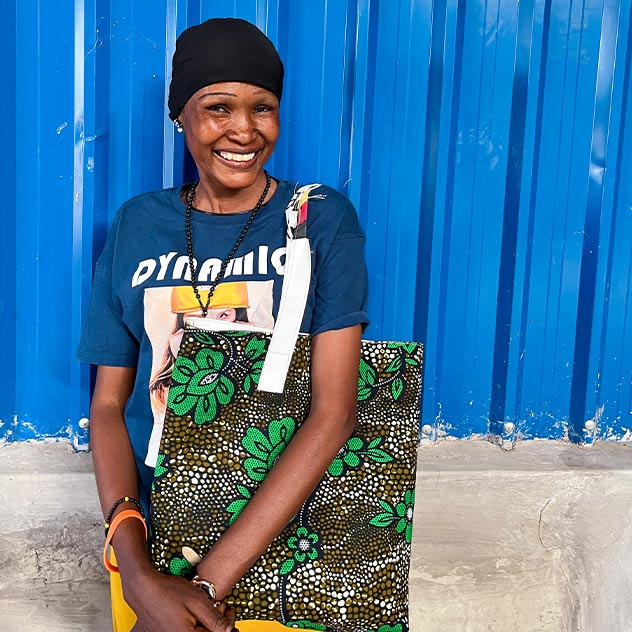 Eine lächelnde Frau steht vor einer blauen Metallwand, trägt ein bedrucktes T-Shirt und eine bunte Stofftasche mit Blumenmuster.