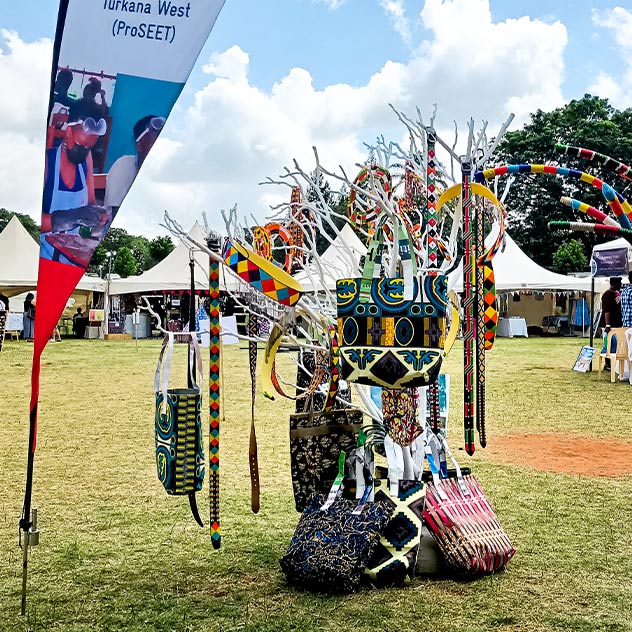 Ein Ausstellungsstand im Freien mit einem Ständer voller bunter Taschen aus Recyclingmaterialien neben einem Banner des Projekts „ProSEET“.