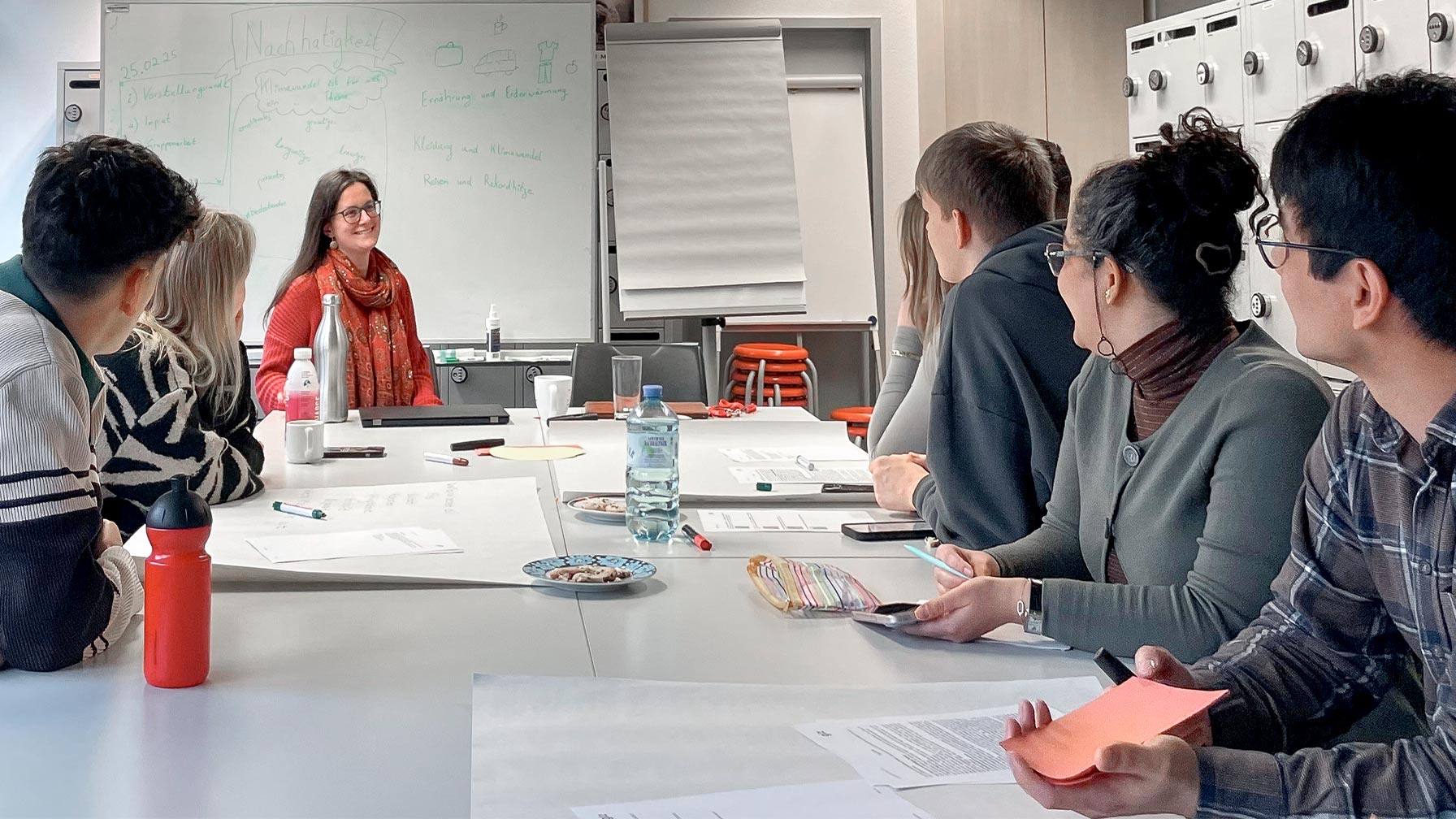 Eine Gruppe von Menschen sitzt in einem Seminarraum an einem langen Tisch, während eine Frau vorne an der Tafel spricht; auf der Tafel steht „Nachhaltigkeit“.