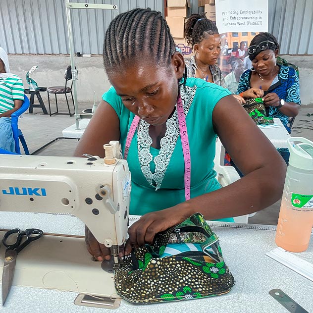 Eine Frau näht konzentriert mit einer Nähmaschine eine Tasche aus Recyclingmaterial, im Hintergrund arbeiten weitere Frauen an ähnlichen Projekten.