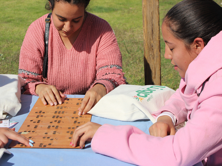 Drei junge Frauen sitzen an einem Tisch im Freien und ertasten mit den Fingern ein hölzernes Brett mit Brailleschrift.