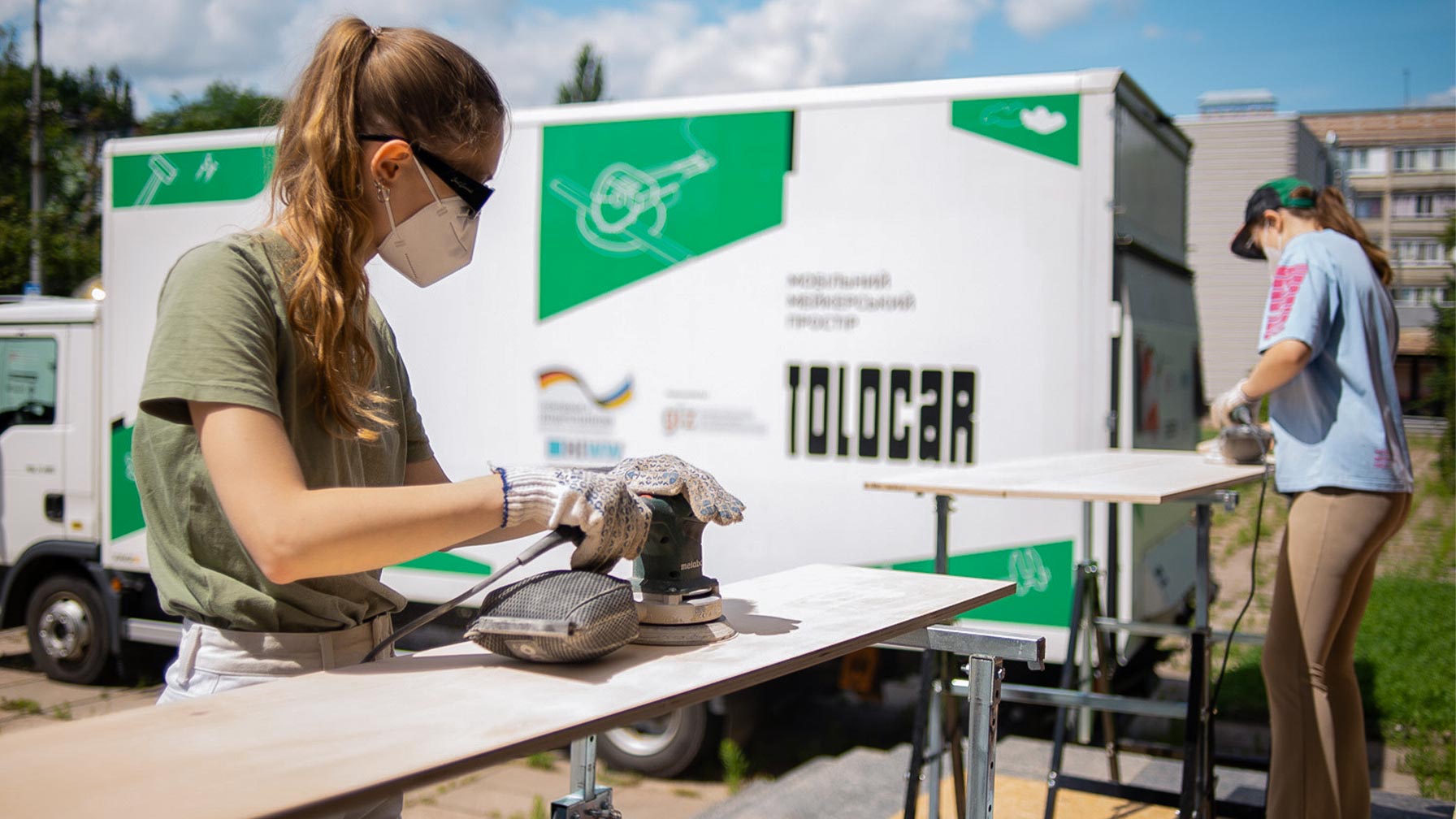 Zwei junge Frauen schleifen mit Schutzmasken und Schleifgeräten Holzbretter im Freien vor einem mobilen Werkstattfahrzeug mit der Aufschrift „Tolocar“.