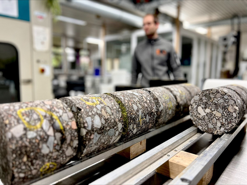 Zylindrisch gebohrte Asphaltproben liegen in Reihe auf einer Metallvorrichtung, im Hintergrund eine Person in einem Labor.