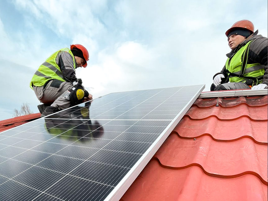 Zwei Personen in Schutzkleidung und Helmen installieren ein Solarpanel auf einem roten Ziegeldach unter bewölktem Himmel.