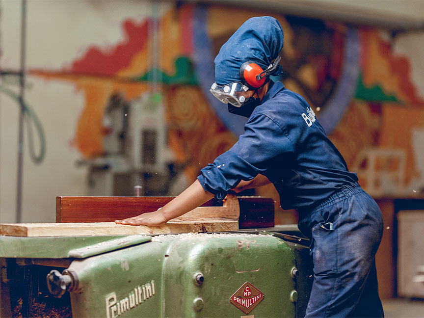 Eine Frau in Arbeitskleidung und Schutzausrüstung bedient eine Holzbearbeitungsmaschine in einer Werkstatt mit bunter Wandmalerei.