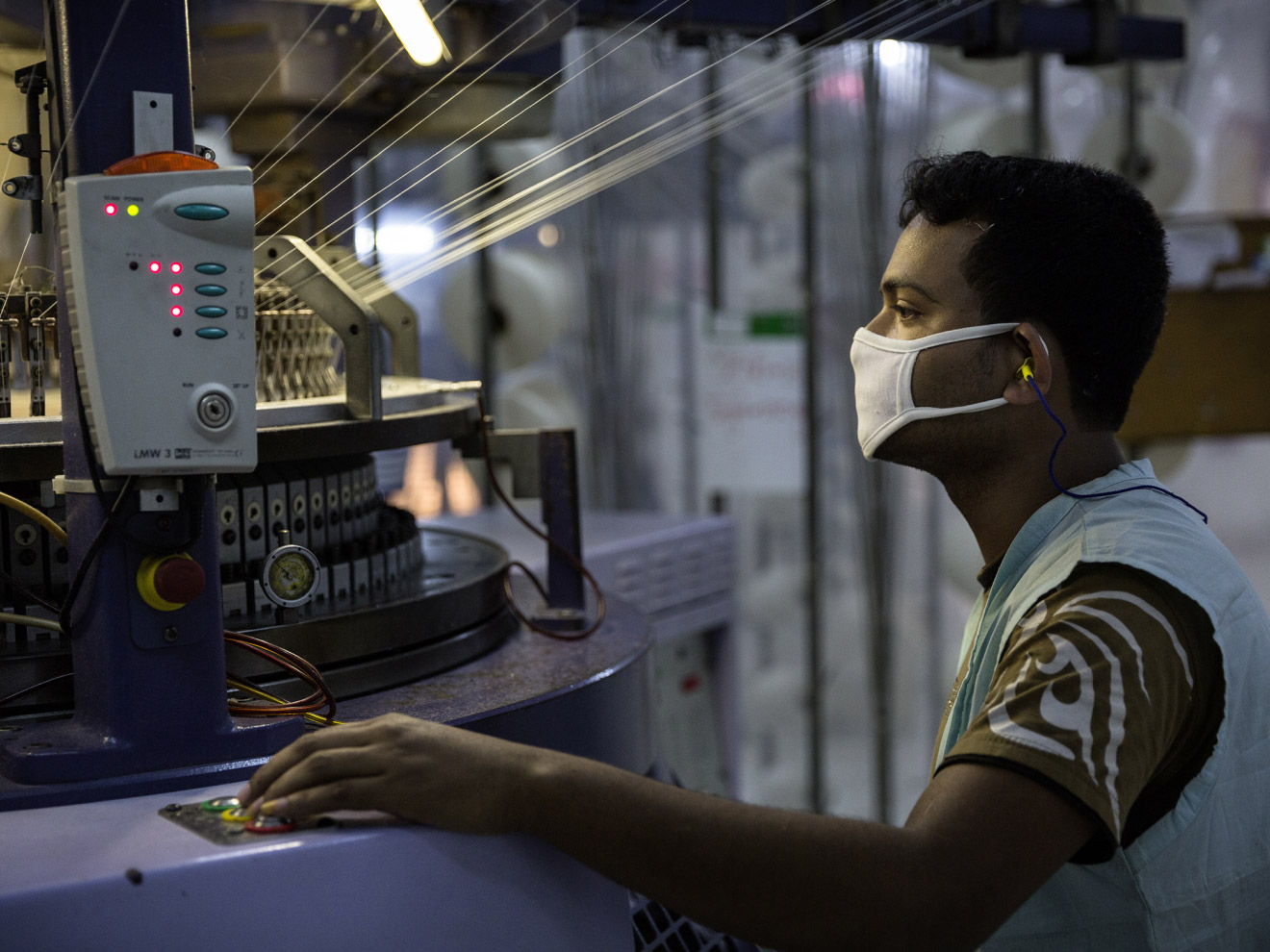 Ein Mann mit Maske und Gehörschutz bedient eine industrielle Textilmaschine und beobachtet aufmerksam den Produktionsprozess.